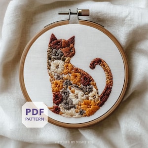 以下が含まれることがあります： 茶色、オレンジ、クリーム色の花柄刺繍が施された猫のシルエットが特徴の刺繍枠。木製の枠が白い布にデザインを固定しています。「PDF PATTERN」の文字が見えます。