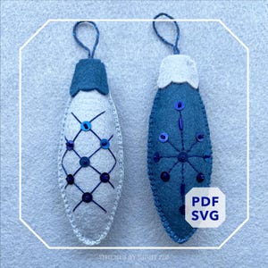 Può includere: Due decorazioni natalizie fatte a mano in feltro. Uno è bianco con un motivo a griglia blu e paillettes, l'altro è blu con un disegno a fiocco di neve e paillettes. Entrambi hanno anelli blu e sono decorati con paillettes.