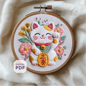 以下が含まれることがあります： 木製のフレームに入った刺繍の招き猫。ピンクと黄色のアクセントが付いた白い猫は、花と緑に囲まれています。猫は片方の足を上げており、日本語の文字が入った金貨を持っています。「DIGITAL PDF」の文字が白い箱に入っています。