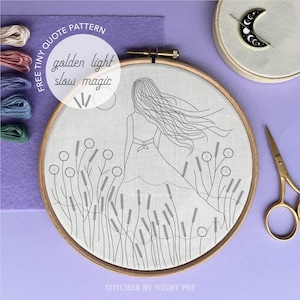 May include: An embroidery hoop with a line drawing of a woman in a field, with the words "golden light slow magic." The hoop is surrounded by embroidery floss, a crescent moon pin, and gold scissors. The background is purple.