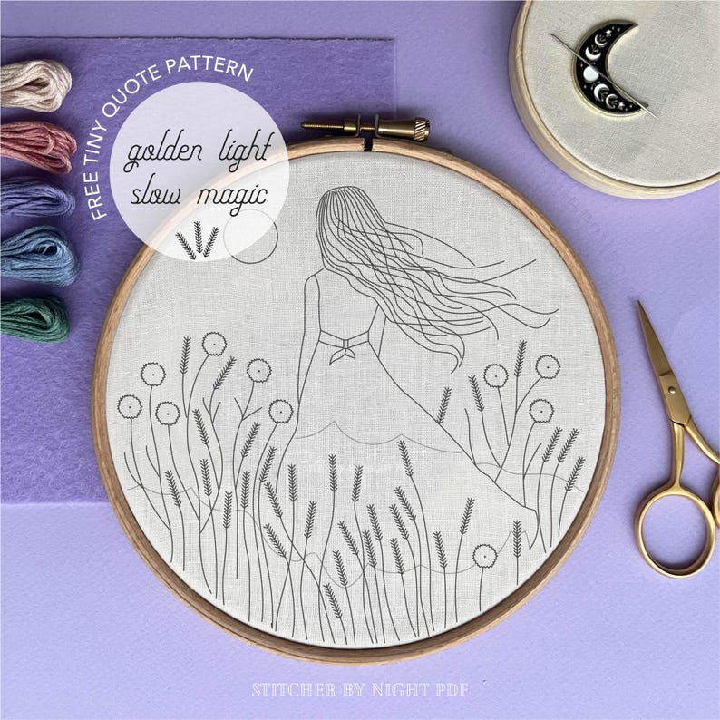May include: An embroidery hoop with a line drawing of a woman in a field, with the words "golden light slow magic." The hoop is surrounded by embroidery floss, a crescent moon pin, and gold scissors. The background is purple.
