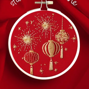 Puede incluir: Aro de bordado rojo con hilo dorado que representa fuegos artificiales y tres linternas colgantes. El texto "stitch good fortune" est&aacute; arqueado sobre el dise&ntilde;o. El texto "MODERN HAND EMBROIDERY PATTERN AVAILABLE NOW ON ETSY" est&aacute; debajo.