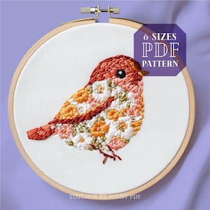 Peut inclure: Un motif de broderie florale d'un oiseau à tête rouge et un corps composé de fleurs colorées. Le motif est affiché sur un tissu blanc dans un cerceau de broderie en bois. Le texte "6 SIZES PDF PATTERN" est affiché dans un hexagone au-dessus du cerceau.