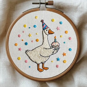 May include: Embroidered hoop art featuring a goose wearing a party hat and holding a cupcake with a lit candle. The design includes colorful stars and dots, with the text "BIRTHDAY GOOSE" and "stitch silly joy".