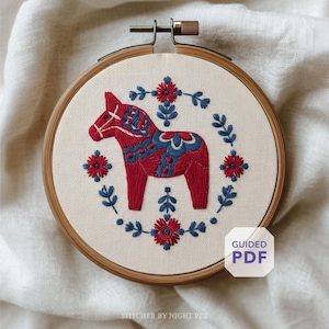 May include: An embroidered Dala horse in a wooden hoop. The horse is red with blue detailing, encircled by a wreath of blue leaves and red flowers. The text "GUIDED PDF" is in a white box. The text "STITCHER BY NIGHT PDF" is at the bottom.
