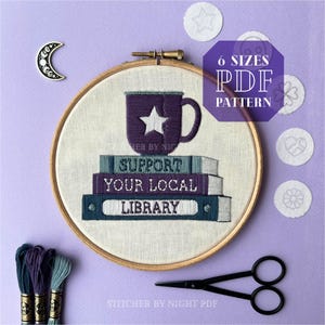 May include: A purple coffee mug with a white star sits on top of a stack of books that say "Support Your Local Library". The books are in a wooden embroidery hoop. The image is on a purple background.