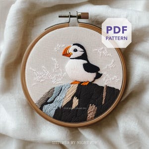 Peut inclure: Broderie d'un macareux moine dans un cercle en bois. L'oiseau a un corps noir et blanc, un bec orange et est perché sur une formation rocheuse. Le texte "PDF PATTERN" est dans un octogone blanc. Le texte "STITCHER BY NIGHT PDF" est en bas.