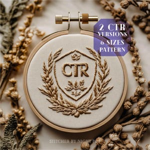 May include: An embroidery hoop with a beige fabric featuring a detailed, gold-colored embroidered design. The design includes the letters "CTR" within a shield shape, surrounded by a wreath. The text "2 CTR VERSIONS 6 SIZES PATTERN" is visible. The hoop is surrounded by dried plants.