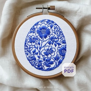Puede incluir: Diseño de bordado floral azul en forma de huevo, en un bastidor de bordado de madera. El diseño presenta flores y hojas detalladas sobre un fondo blanco. El texto "GUIDED PDF" está en la esquina inferior derecha.