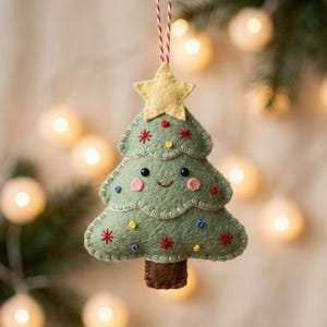 May include: A felt Christmas tree ornament in sage green with a yellow star on top and a brown trunk. The tree features embroidered red starbursts, blue and yellow dots, pink cheeks, and black eyes, hanging from a red and white string.