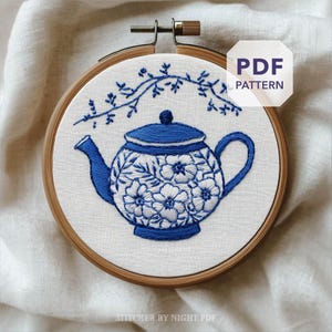 Può includere: Ricamo di un disegno di teiera in un telaio di legno. La teiera è blu e bianca, con dettagli floreali. Un ramo floreale blu è sopra la teiera. Il testo "PDF PATTERN" è in una casella bianca. Il testo "STITCHER BY NIGHT PDF" è in basso.