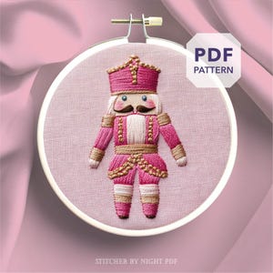 May include: Embroidered pink and gold nutcracker design in a round embroidery hoop. The nutcracker has a pink hat, uniform, and legs, with gold accents and a white face and mustache. The text "PDF PATTERN" is visible.