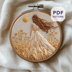 Puede incluir: Bordado en bastidor que representa a una mujer con un vestido blanco en un campo de trigo, con un sol amarillo. El bordado utiliza hilos amarillos y marrones. El texto "PDF PATTERN" es visible.
