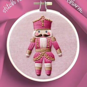 May include: An embroidery hoop featuring a pink and gold embroidered nutcracker soldier. The soldier has a pink uniform, a white beard, and a mustache. The text "stitch nucracker soldier charm" is arched above the hoop. The text "MODERN HAND EMBROIDERY INSTANT DOWNLOAD" is below the hoop.