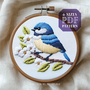 May include: Embroidered design of a bluebird perched on a branch with white blossoms. The design is framed in a wooden embroidery hoop. The text "6 SIZES PDF PATTERN" is in a purple box above the hoop. The text "STITCHER BY NIGHT PDF" is below the hoop.
