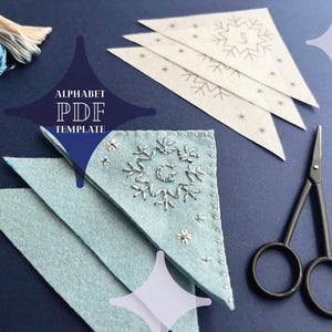 May include: A set of three white felt snowflake templates with a black outline and a letter J in the center of each snowflake. The templates are stacked on top of each other. There is also a blue felt snowflake with white embroidery of a snowflake and a letter G in the center. A pair of black embroidery scissors are on the right side of the image. The text "ALPHABET PDF TEMPLATE" is in the center of the image.