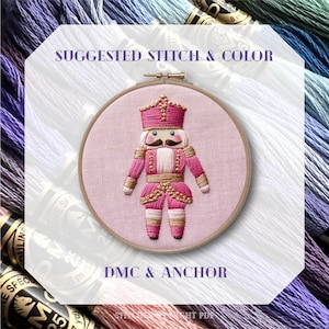 May include: An embroidery hoop featuring a pink and gold nutcracker design. The nutcracker is wearing a pink uniform with gold accents and a crown. The text "SUGGESTED STITCH & COLOR" and "DMC & ANCHOR" are visible. The background includes spools of thread.