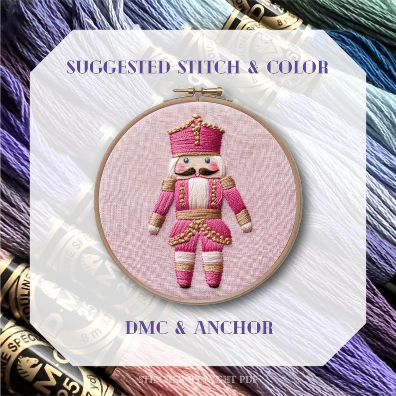 May include: An embroidery hoop featuring a pink and gold nutcracker design. The nutcracker is wearing a pink uniform with gold accents and a crown. The text "SUGGESTED STITCH & COLOR" and "DMC & ANCHOR" are visible. The background includes spools of thread.