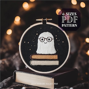 Op de afbeelding: Een zwarte borduurring met een witte geest die een bril draagt en op een stapel boeken zit. De achtergrond is donkerblauw met gouden sterren. De tekst "6 SIZES PDF PATTERN" staat in een witte doos met een zwarte rand.