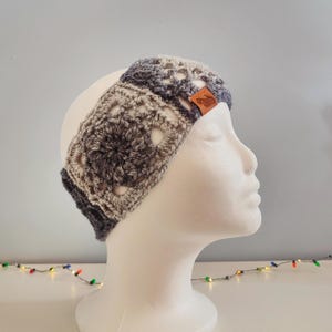 May include: Gray and beige crocheted headband with a brown leather label. The headband is on a white mannequin head.