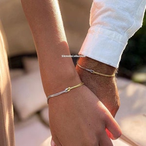 May include: Two matching bracelets with a knot design. One bracelet is silver and the other is gold. The bracelets are worn on the wrists of two people holding hands.