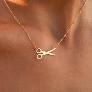 May include: A gold-coloured necklace with a scissors pendant. The scissors are detailed and suspended from a delicate chain. The necklace is worn around the neck, against a skin tone background.