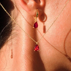 Op de afbeelding: Een gouden ketting met een traanvormige rode edelsteen hanger. De ketting wordt gedragen met een paar gouden oorringen met een bijpassende rode edelsteen hanger.