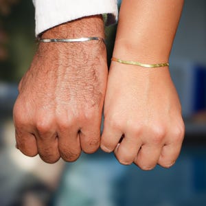 May include: Two hands, one with a silver bracelet and the other with a gold bracelet. The bracelets have a simple, wavy design. The background is blurred, with hints of blue and green.