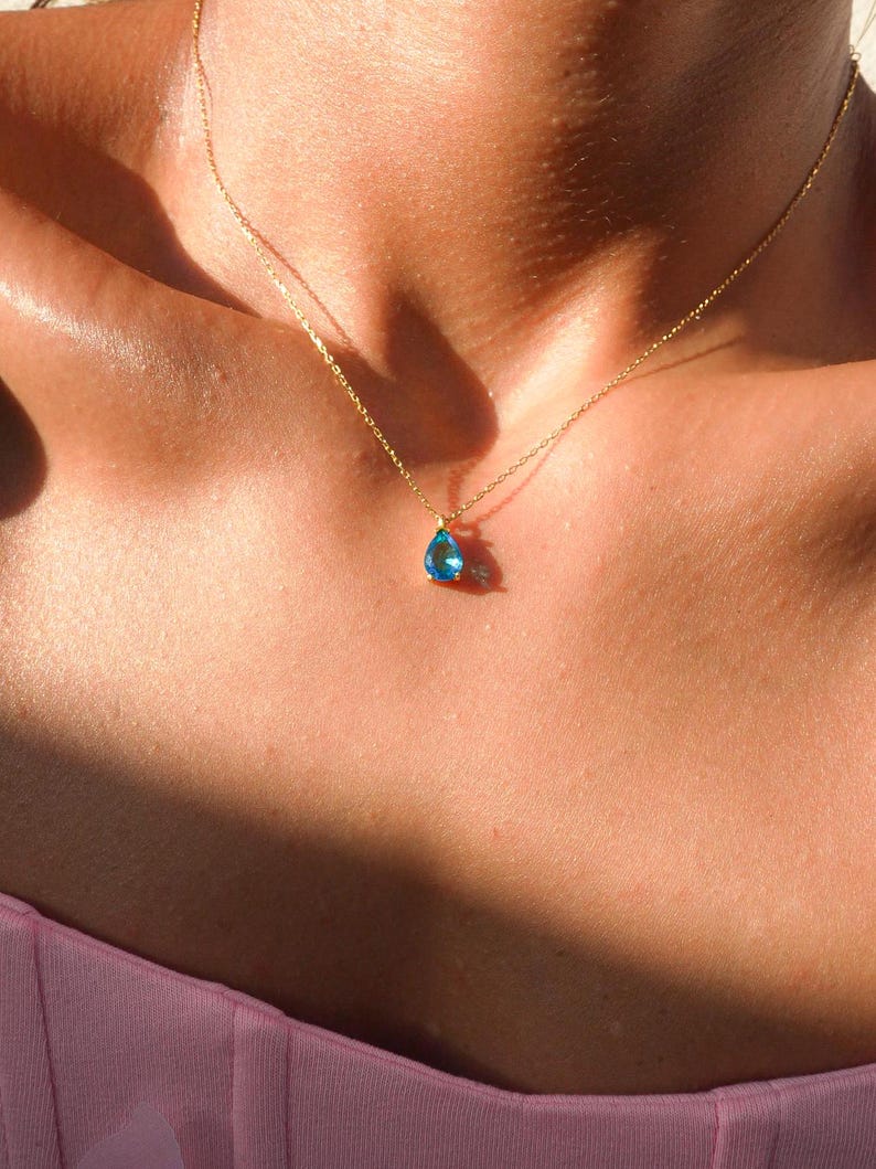 May include: A delicate gold chain necklace with a teardrop-shaped blue gemstone pendant.