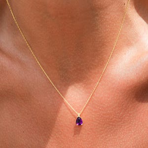 May include: A delicate gold chain necklace with a small, teardrop-shaped purple gemstone pendant.