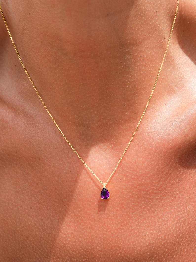 May include: A delicate gold chain necklace with a small, teardrop-shaped purple gemstone pendant.
