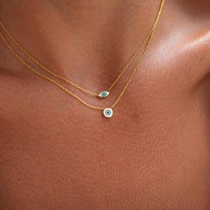 May include: Two gold necklaces with blue evil eye pendants. The top necklace has a smaller pendant and the bottom necklace has a larger pendant.