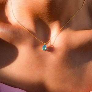 Pode incluir: Um delicado colar de corrente de ouro com um pequeno pingente de pedra preciosa azul em forma de lágrima.