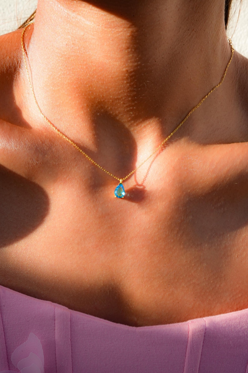 May include: A delicate gold chain necklace with a small teardrop-shaped blue gemstone pendant.