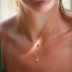 May include: A woman wearing a green top with two gold necklaces. The top necklace has a green gemstone pendant and the bottom necklace has a yellow gemstone pendant.