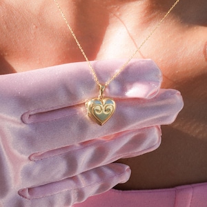 Op de afbeelding: Een gouden hartvormige hanger aan een delicate gouden ketting. De hanger heeft een gedetailleerd ontwerp en wordt gedragen door een persoon met een roze top.