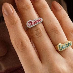 May include: Two gold rings with the names "Claire" and "Sean" spelled out in cursive script. The rings are set with small clear stones and have a red and green enamel finish.