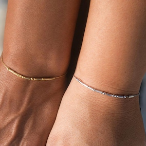 May include: Two delicate bracelets worn on wrists, one in a gold tone and one in a silver tone. Each bracelet features a thin chain adorned with a series of small, cylindrical and spherical metal beads arranged in a pattern resembling Morse code.