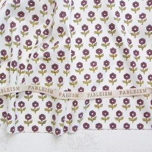 May include: A white fabric featuring a repeating pattern of small purple flowers with green stems and leaves. A beige ribbon runs horizontally across the fabric, printed with the repeated text FABLEISM in a dark brown sans-serif font.