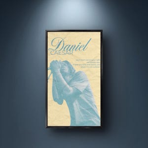 May include: A framed poster featuring singer Daniel Caesar in a light blue hue against a beige background. The poster includes the singer's name and song lyrics in a stylized font. The frame is black, and the poster is mounted on a dark blue wall.