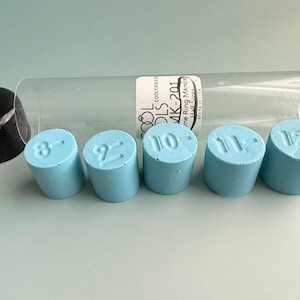 May include: Five cylindrical light blue ring sizing inserts with embossed numbers 8, 9, 10, 11, and 12, arranged in a row. A clear plastic storage tube with a black cap and a label reading COOL TOOLS MK-201 is positioned behind the inserts.