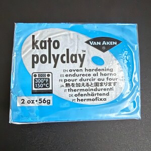 May include: A package of Kato Polyclay in a blue wrapper. The package includes the text "kato polyclay" in white, along with the brand name "VAN AKEN". It also indicates the clay is oven hardening at 300°F (150°C) and contains 2 oz (56g).