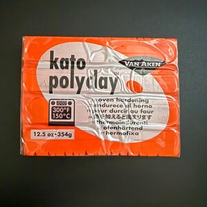 May include: A package of Kato PolyClay in an orange wrapper. The package has the text "kato polyclay" in black and white, along with temperature and weight information. The package weighs 12.5 oz (354g).