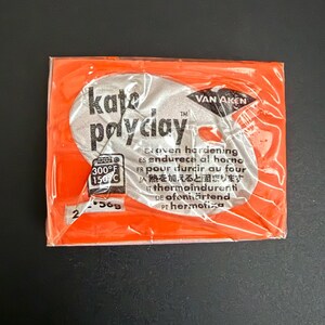 May include: An orange package of Kate Payclay oven-hardening clay. The package has a silver oval with the brand name and text in multiple languages. The package also shows the temperature in Fahrenheit and Celsius.