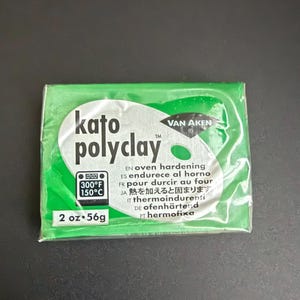 May include: A package of Kato Polyclay in a green wrapper. The package includes the text "kato polyclay" and "2 oz • 56g". It also includes the oven temperature in Fahrenheit and Celsius.