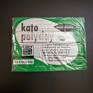 May include: A package of Kato Polyclay, an oven-hardening polymer clay. The package is wrapped in clear plastic with a green and silver label. The label includes the brand name, temperature settings of 300°F (150°C), and the weight of 12.5 oz (354g).