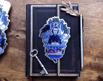 Può includere: Un adesivo con un'illustrazione blu e bianca di Edgar Allan Poe seduto a una scrivania con un corvo, uno scheletro e altre immagini gotiche. L'adesivo è su un diario di pelle nera con una chiave e una foglia.
