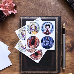 May include: Six round stickers with various illustrations. The stickers feature a woman with long blonde hair holding a pumpkin, a woman with dark hair wearing a red dress, a black cat with a clock, a man with a clock, a black cat with a clock, and a black cat with a clock. The stickers are on a dark brown leather journal.