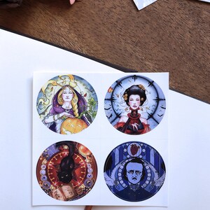May include: Four round stickers with colorful illustrations. The first sticker features a woman in a purple robe holding a pumpkin. The second sticker features a woman in a red dress with a clock behind her. The third sticker features a black cat on a red background with a book and a clock. The fourth sticker features a man with a beard in a blue suit with a heart and a clock behind him.