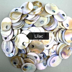 May include: A collection of round, flat shell discs with iridescent surfaces in shades of white, cream, tan, and soft purple. Each disc features a single circular hole near the edge. A black rectangular label with the white text Lilac is centered.